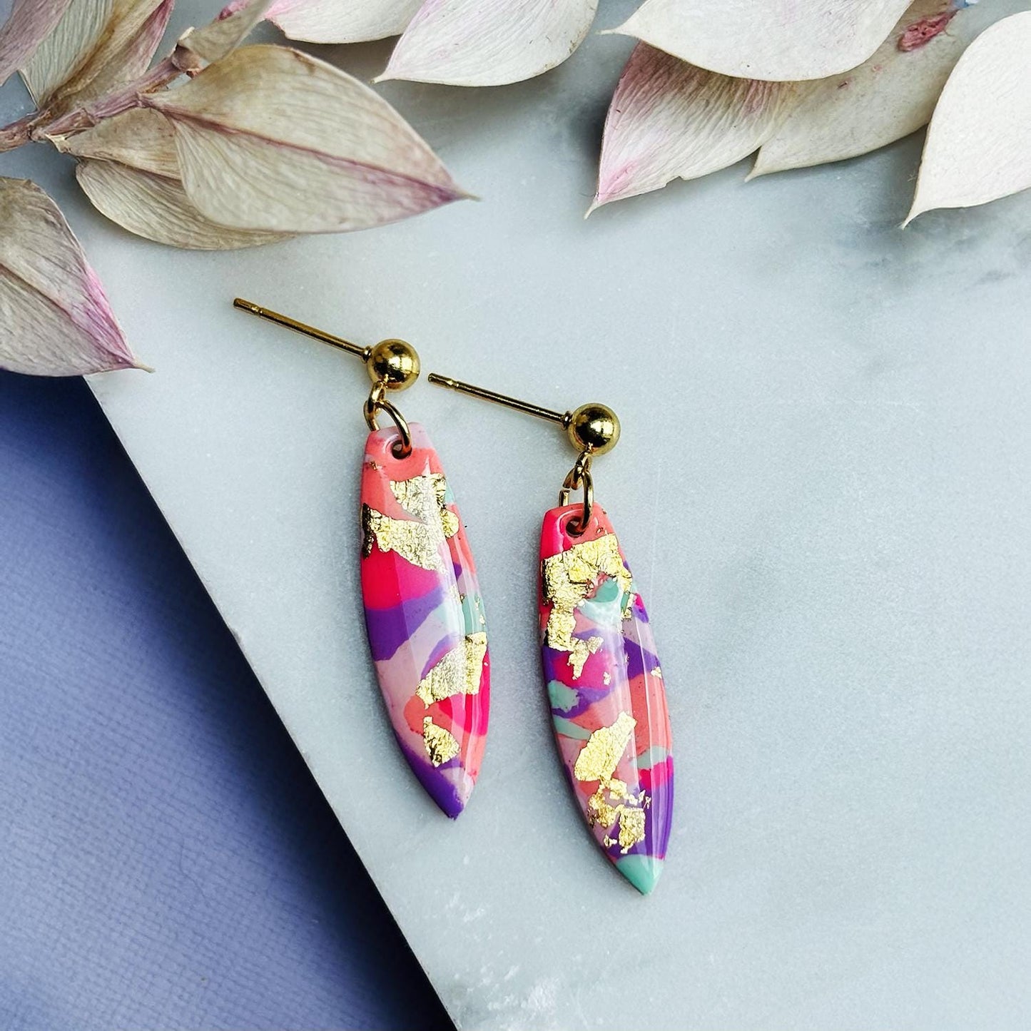 Colourful surfboard-shaped surf earrings with gold foil details, handmade from polymer clay, featuring hypoallergenic surgical stainless steel posts and backings.