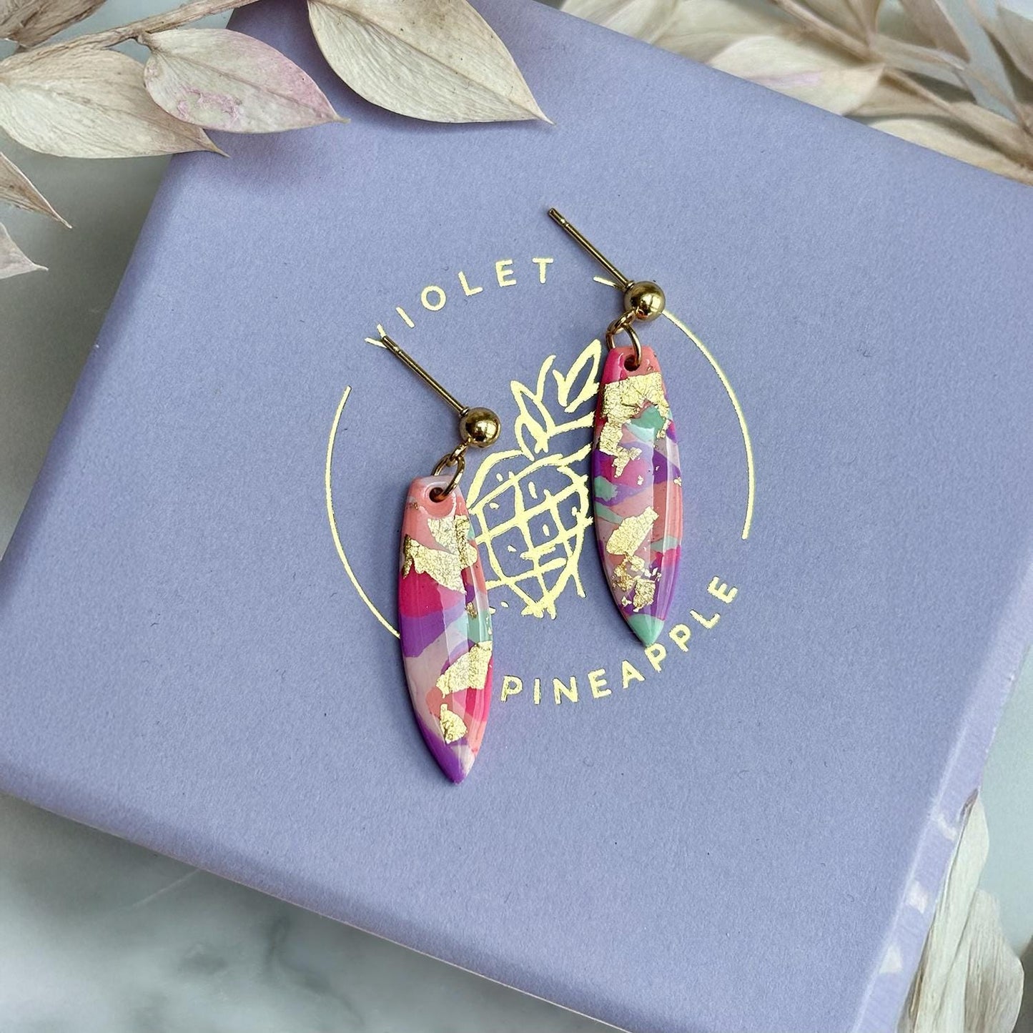 Colourful surfboard-shaped surf earrings with gold foil details, handmade from polymer clay, featuring hypoallergenic surgical stainless steel posts and backings.