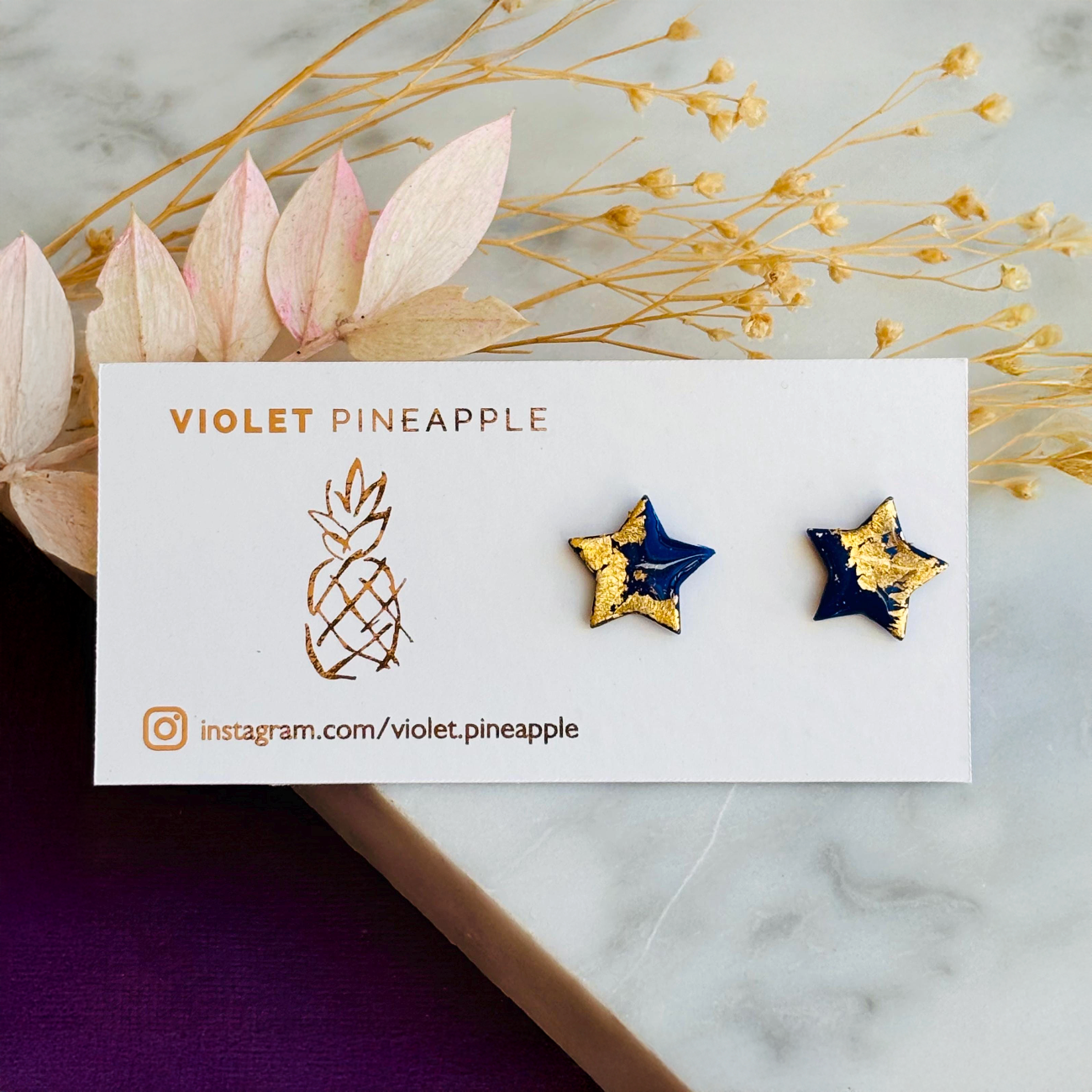 Close-up photograph of a pair of handmade navy blue polymer clay star-shaped stud earrings featuring shimmering gold foil details. The earrings are attached to hypoallergenic surgical steel posts and displayed on a white or neutral background. Minimalist jewelry design.