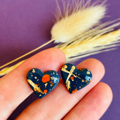 Abstract heart-shaped stud earrings with navy blue, vibrant orange, and gold splatter details, handmade with polymer clay.