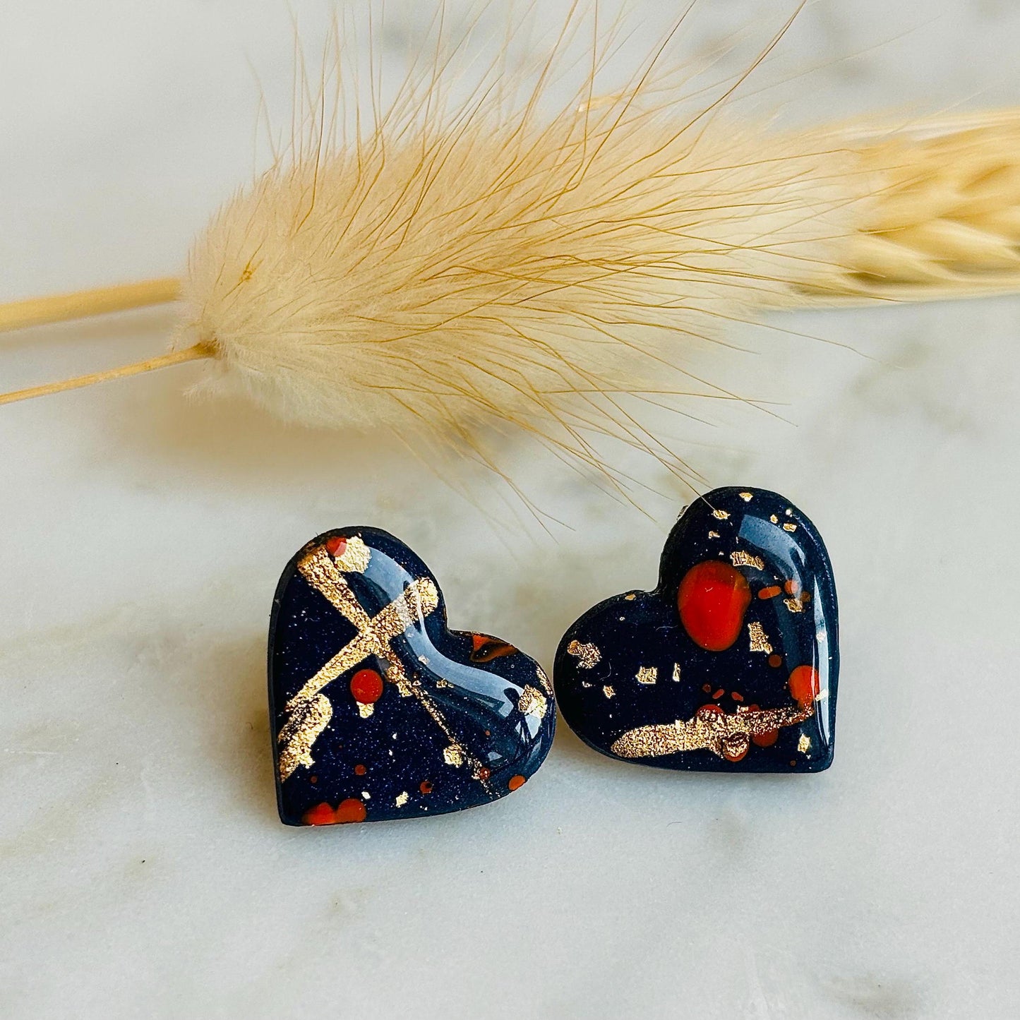 Abstract heart-shaped stud earrings with navy blue, vibrant orange, and gold splatter details, handmade with polymer clay.