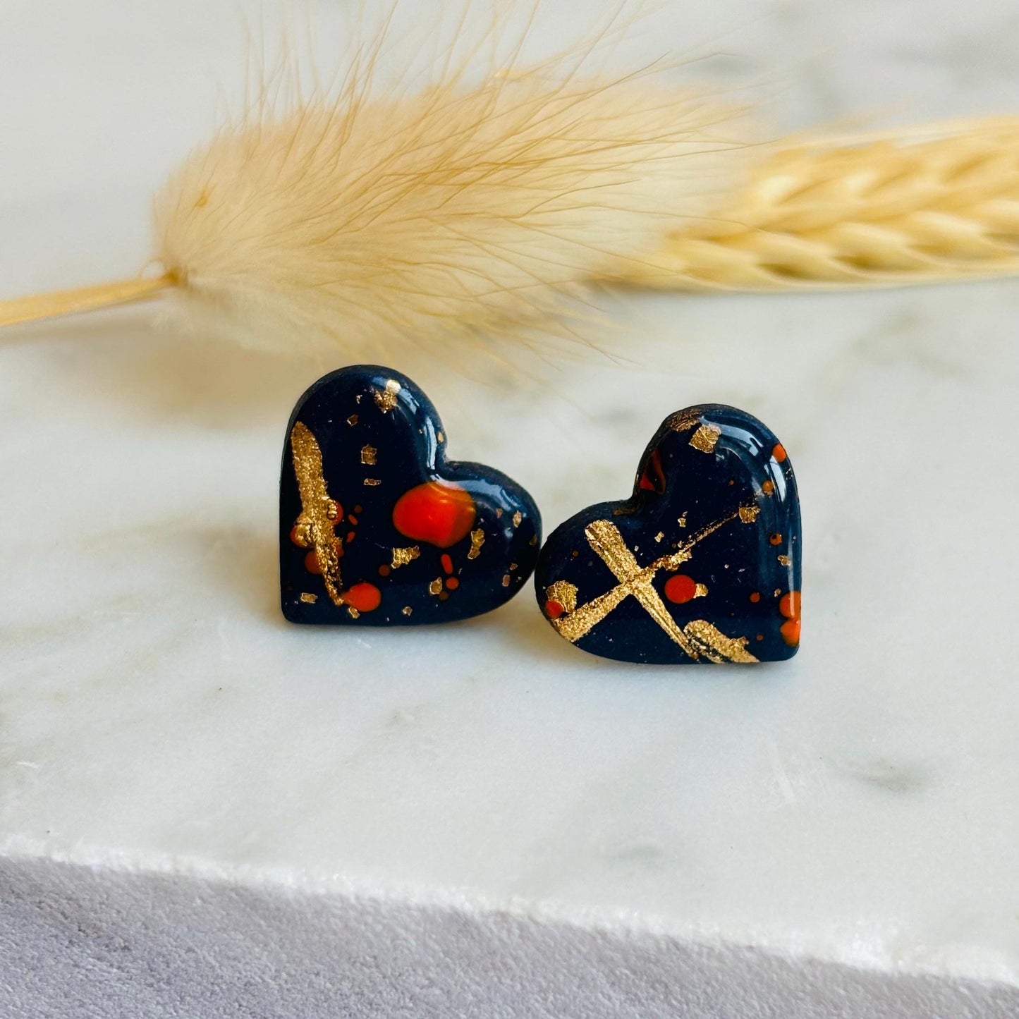 Abstract heart-shaped stud earrings with navy blue, vibrant orange, and gold splatter details, handmade with polymer clay.