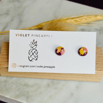 Mini stud earrings with a warm color palette of plum, blush pink, and ochre yellow with gold foil accents, handmade with polymer clay.