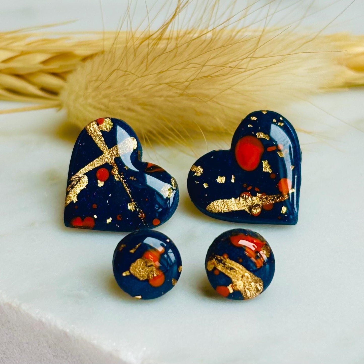 Abstract heart-shaped stud earrings with navy blue, vibrant orange, and gold splatter details, handmade with polymer clay.