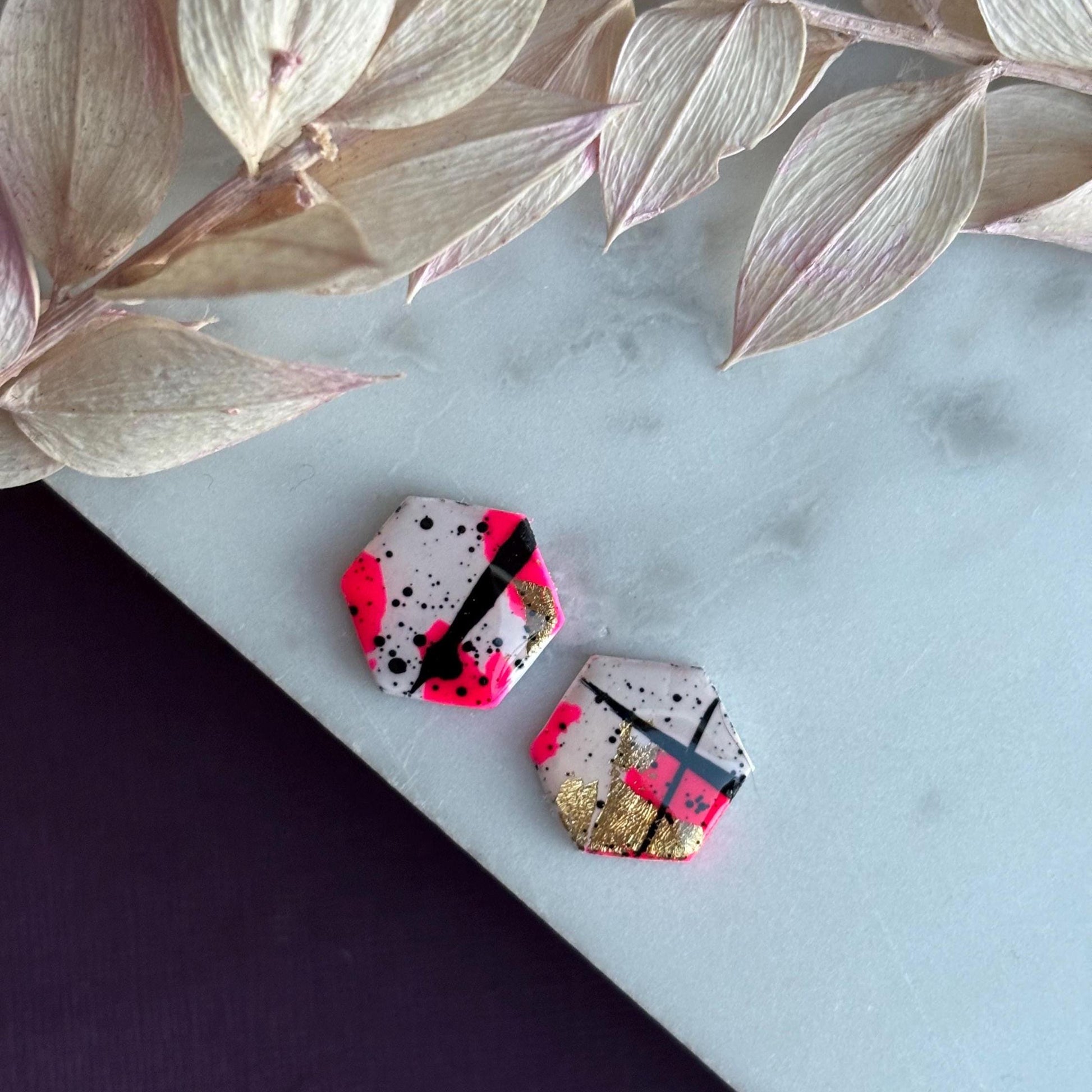 Bold abstract hexagon stud earring featuring white, neon pink, gold foil, and black ink splatter, handmade from polymer clay.