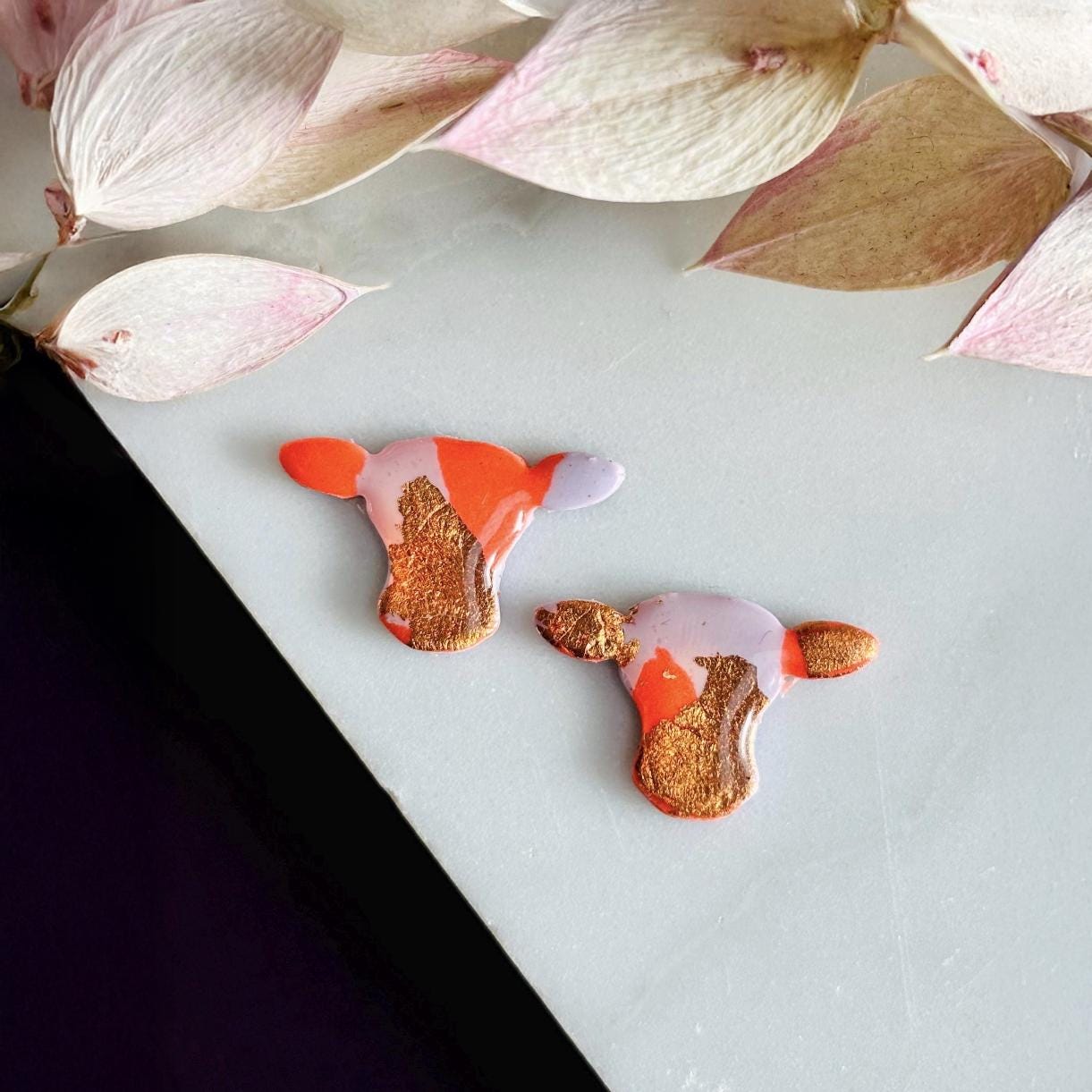 Close-up of small orange and lilac cow stud earrings with gold foil.
