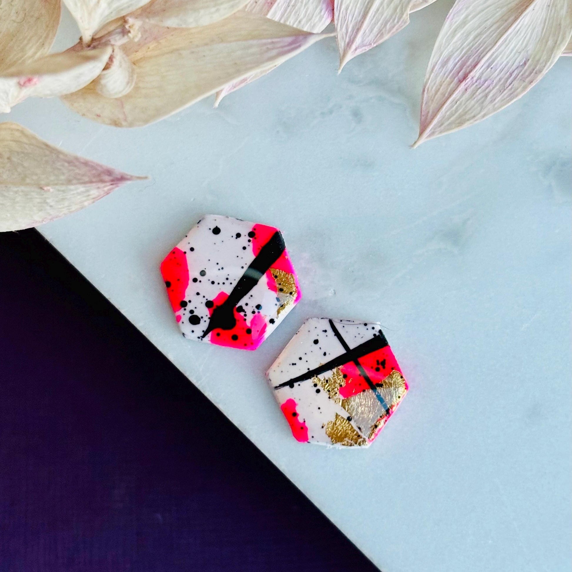 Bold abstract hexagon stud earring featuring white, neon pink, gold foil, and black ink splatter, handmade from polymer clay.