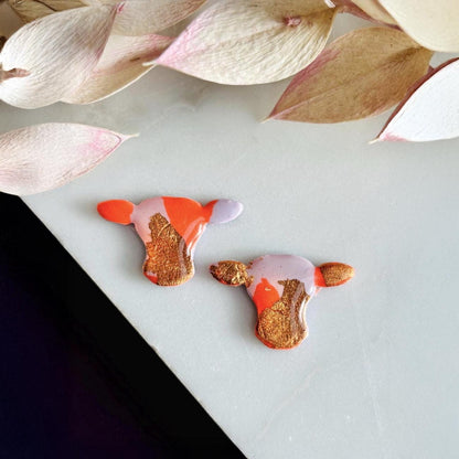 Close-up of small orange and lilac cow stud earrings with gold foil.