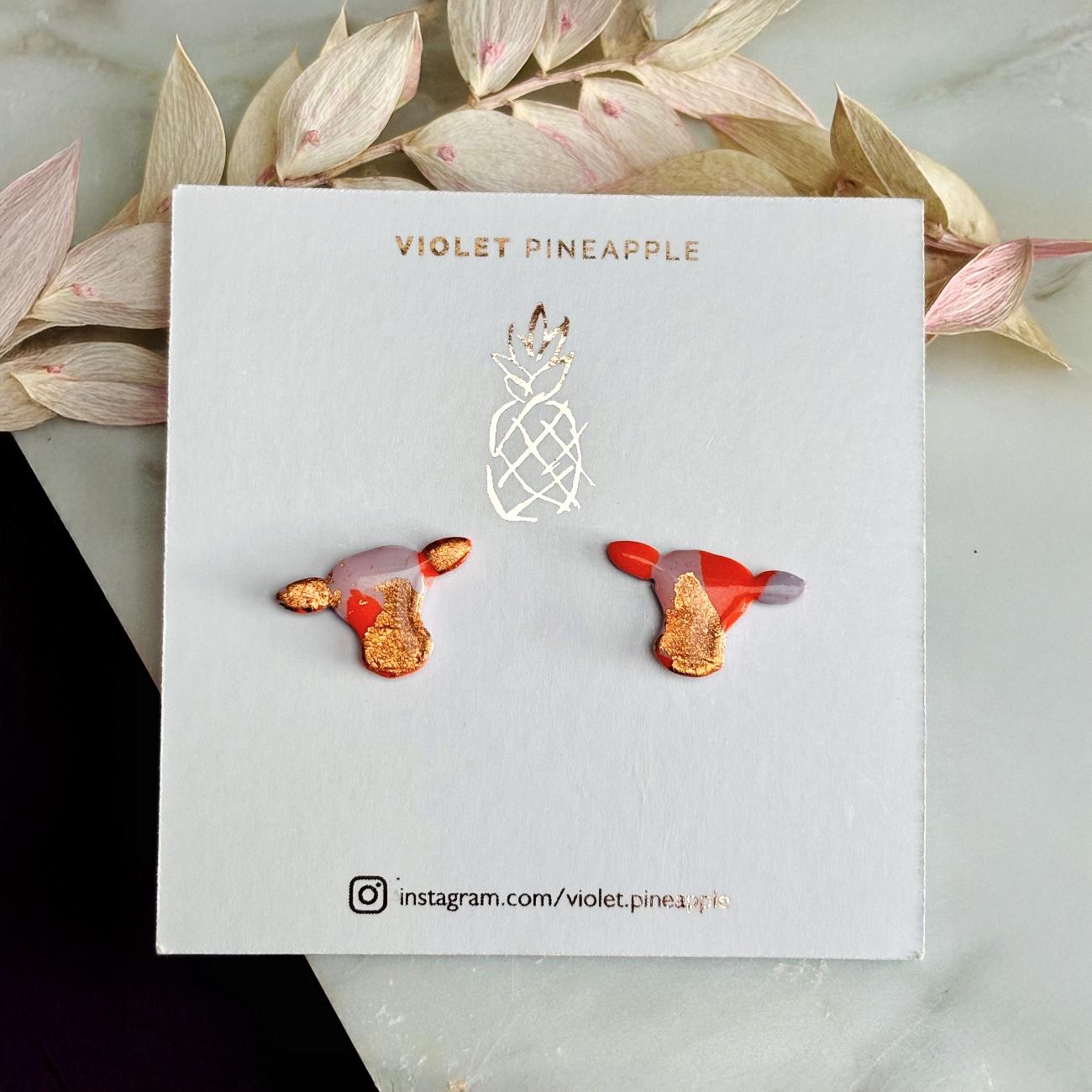 Close-up of small orange and lilac cow stud earrings with gold foil.