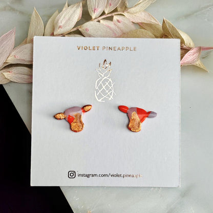 Close-up of small orange and lilac cow stud earrings with gold foil.