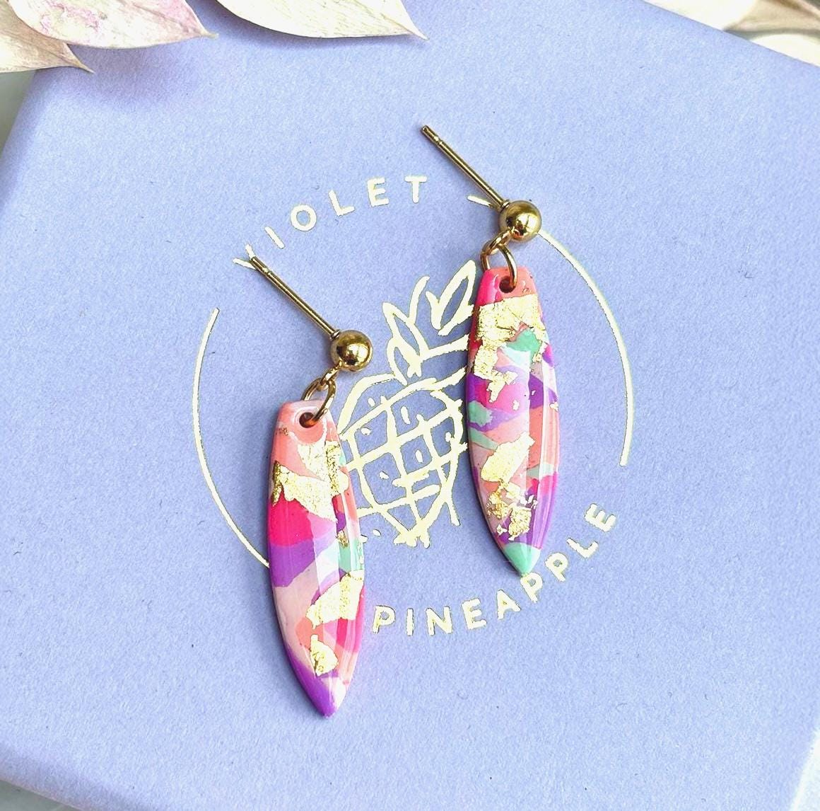Colourful surfboard-shaped surf earrings with gold foil details, handmade from polymer clay, featuring hypoallergenic surgical stainless steel posts and backings.