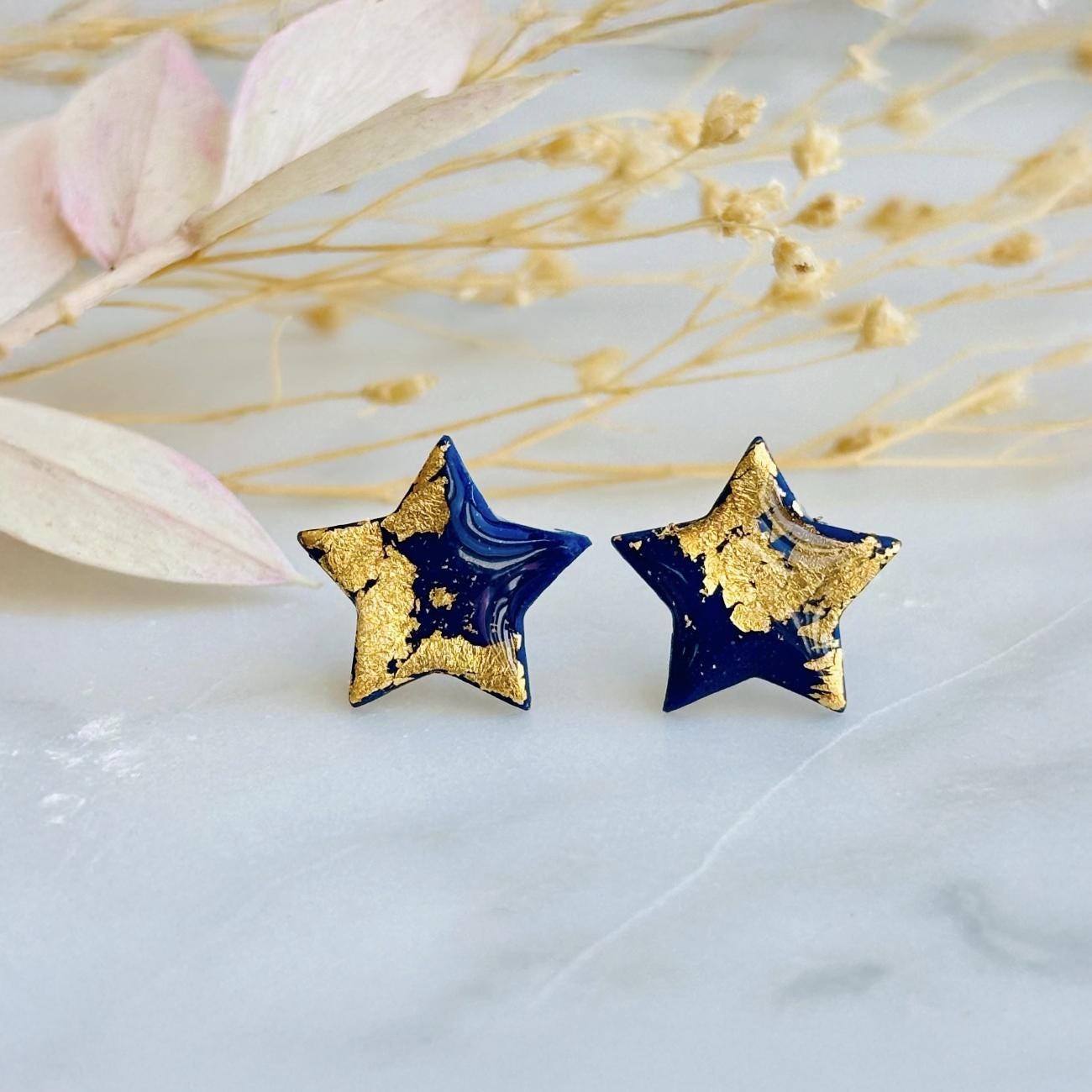 Close-up photograph of a pair of handmade navy blue polymer clay star-shaped stud earrings featuring shimmering gold foil details. The earrings are attached to hypoallergenic surgical steel posts and displayed on a white or neutral background. Minimalist jewelry design.