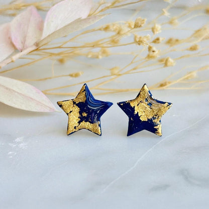 Close-up photograph of a pair of handmade navy blue polymer clay star-shaped stud earrings featuring shimmering gold foil details. The earrings are attached to hypoallergenic surgical steel posts and displayed on a white or neutral background. Minimalist jewelry design.