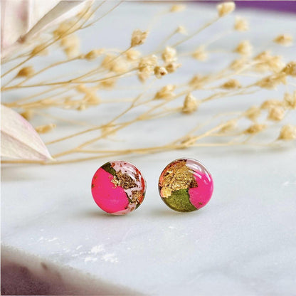 Round earrings with pink and gold design on a white surface with dried flowers.