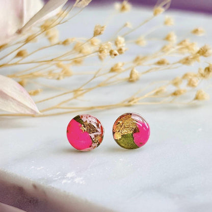 Round earrings with pink and gold design on a white surface with dried flowers.