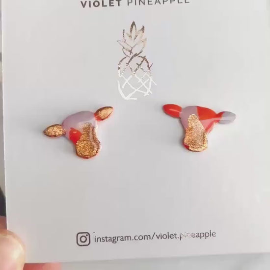 Close-up of small orange and lilac cow stud earrings with gold foil.