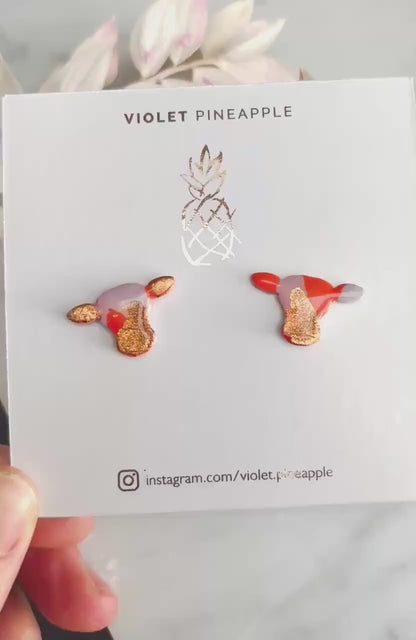 Close-up of small orange and lilac cow stud earrings with gold foil.